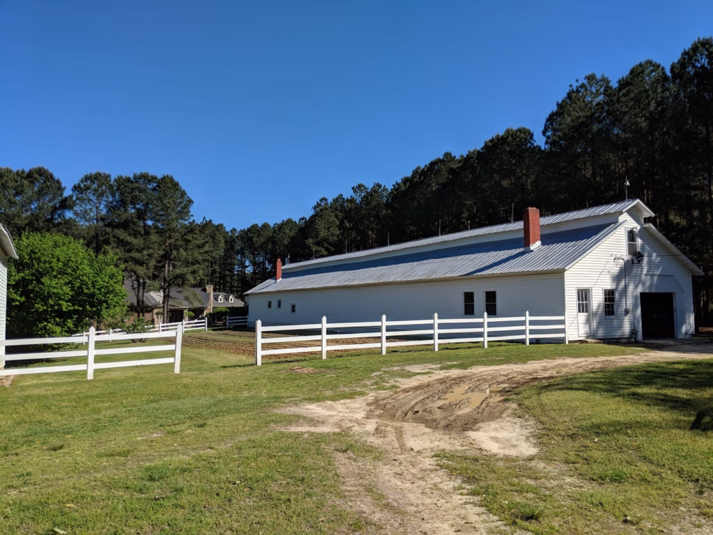 White three-rail farm fence running past a metal-roofed barn on a Zebulon, NC property, installed by Two Brothers Fencing